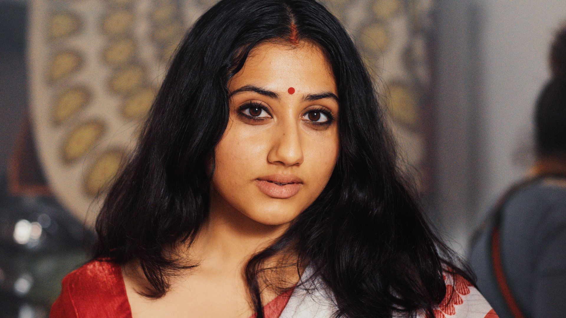 A woman wearing a sari and a red bindi looks into the camera.