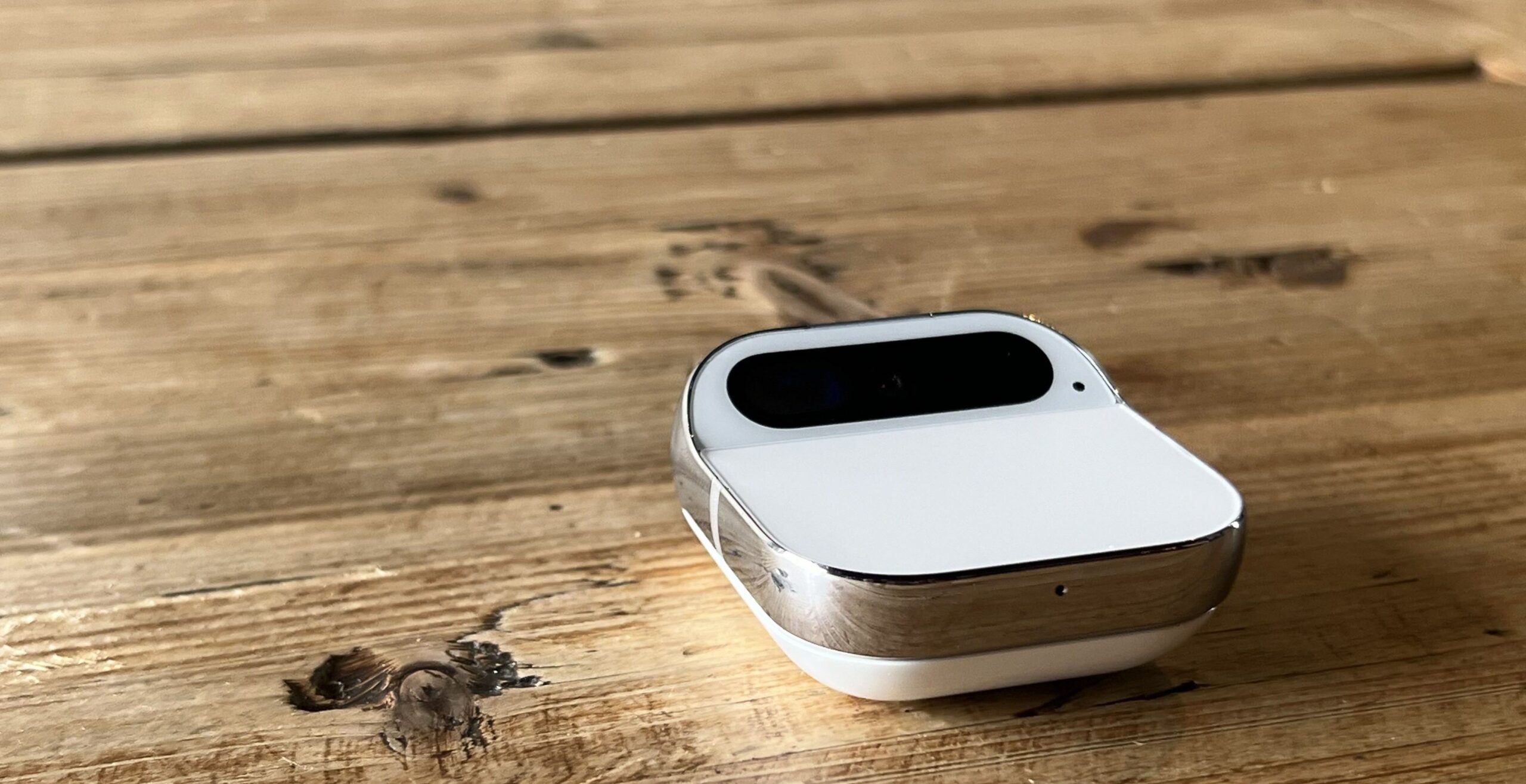  This image features a close-up of a modern, minimalist design earbud case placed on a textured wooden surface. The case is white with a rounded shape and a metallic accent, likely indicating where it opens. The case is partially open, revealing a glimpse of the black interior where the earbuds would be housed. There is a small knot in the wood directly next to the case, adding a rustic element to the sleek technology.