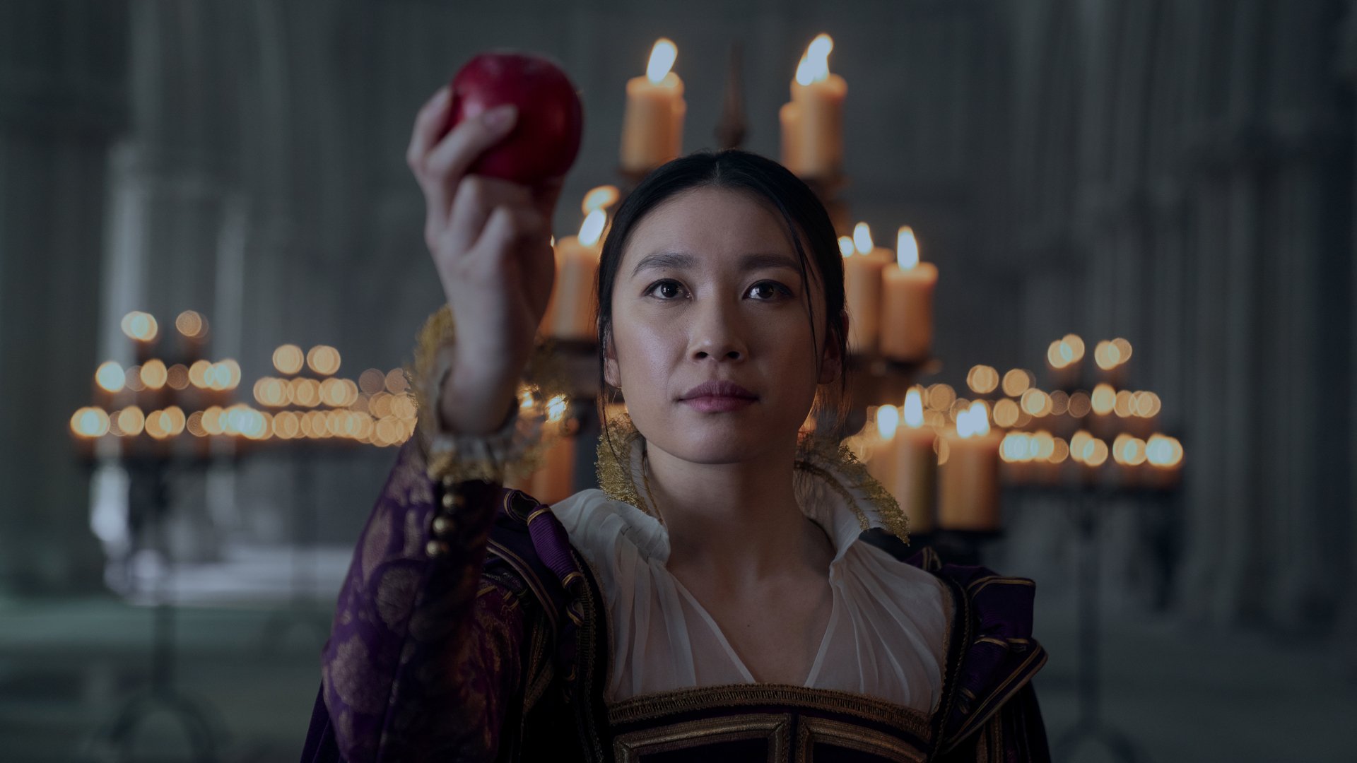 A woman in a 17th century England dress holds out an apple.