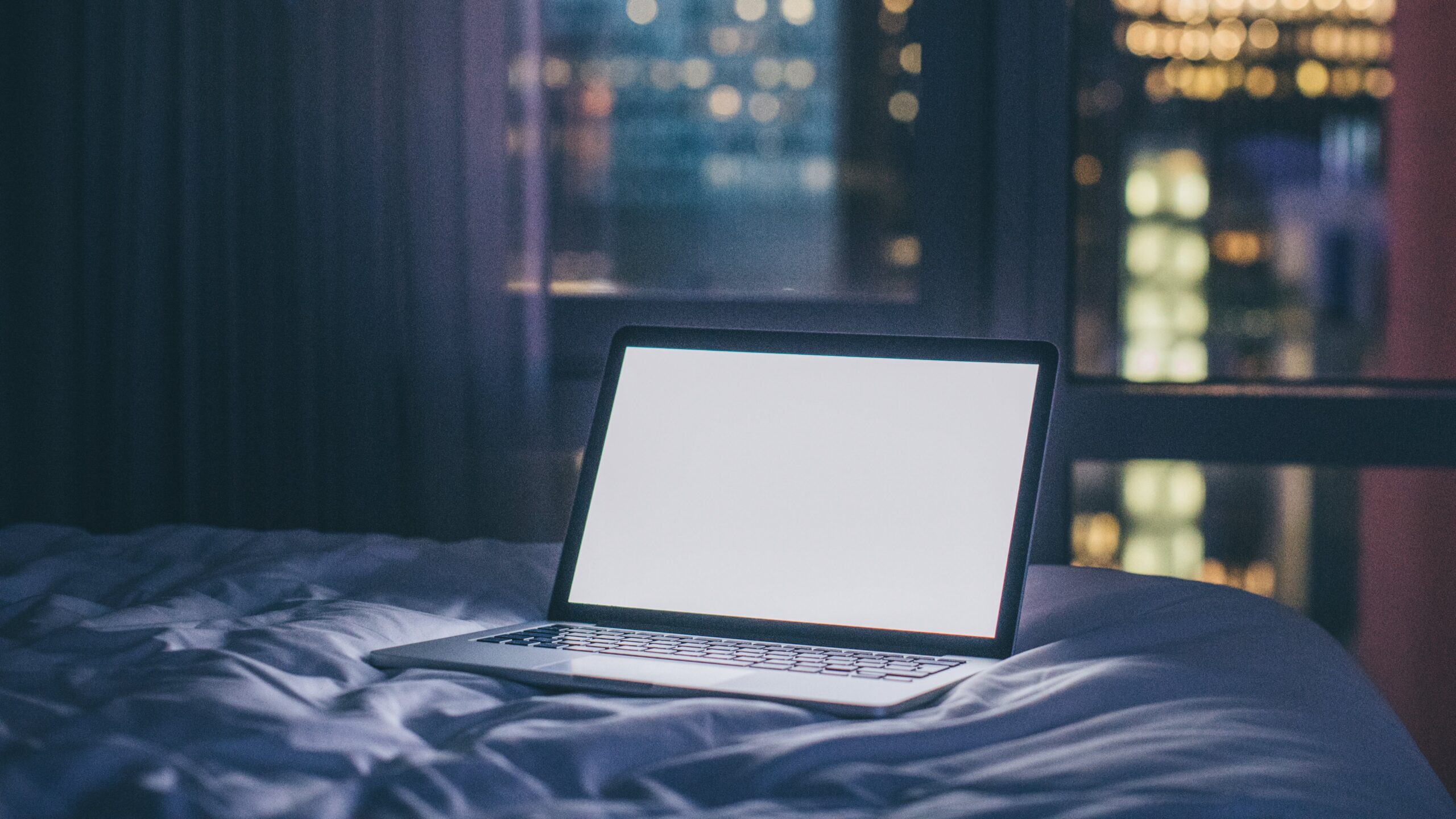 laptop with white screen on bed, city night scene in window