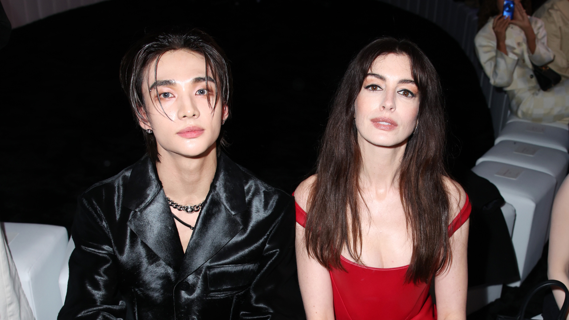 Hyunjin and Anne Hathaway sit side by side. He is in a black leather jacket and she is in a red dress.