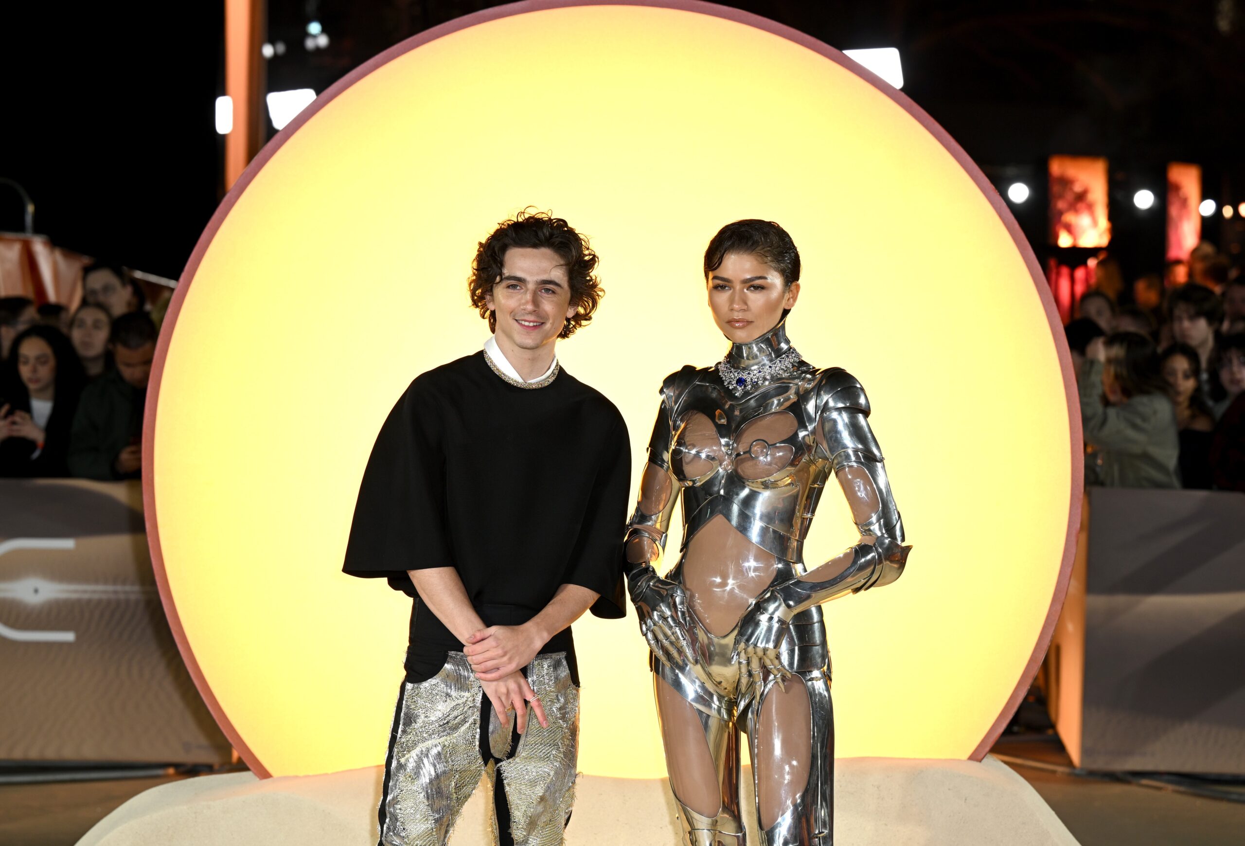 Timothee Chalamet and Zendaya on the 'Dune: Part 2' red carpet illuminated by a glowing yellow circle behind them.