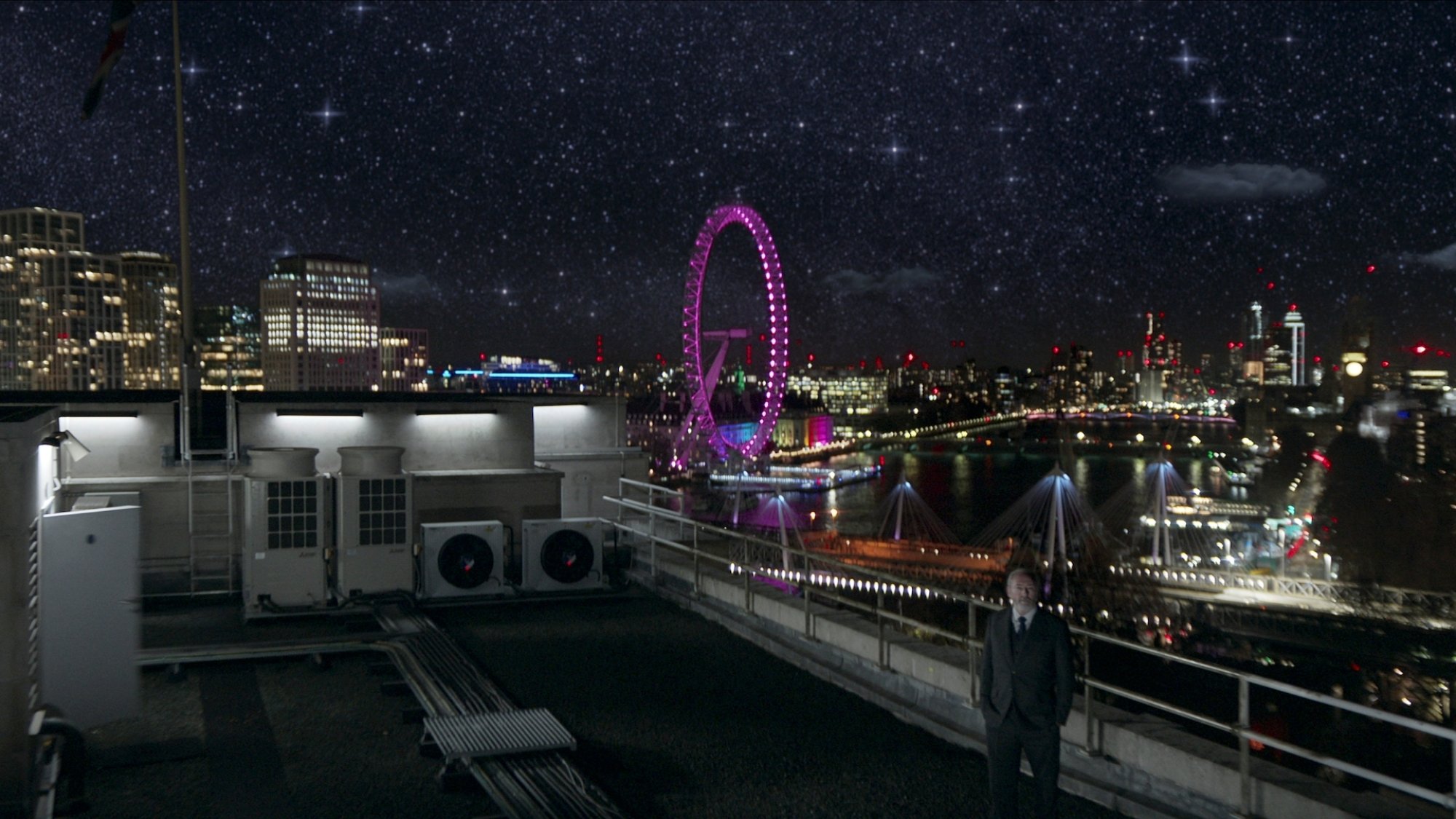 A man stands on a rooftop under the stars with the London skyline in the background.