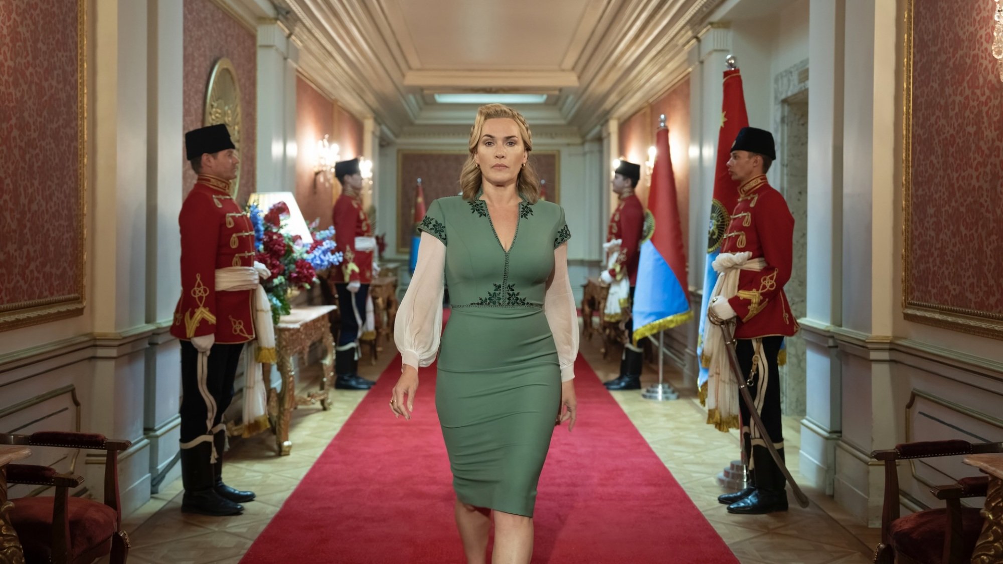 A woman in a green dress with puffy white sleeves walks down a hallway lined with soldiers in red uniforms.