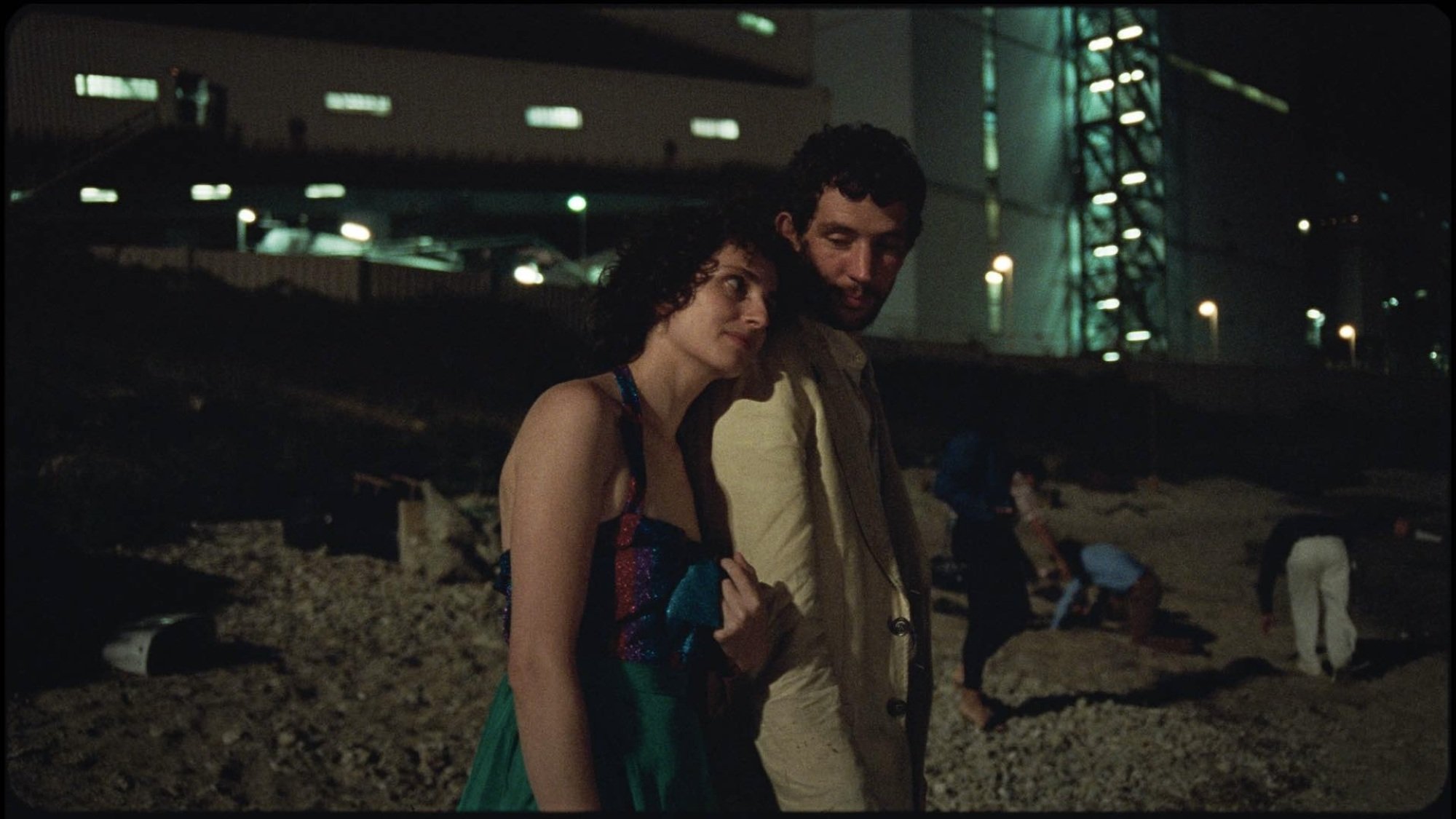 A woman leans her head on a man's shoulder while they stand on a beach below a factory.