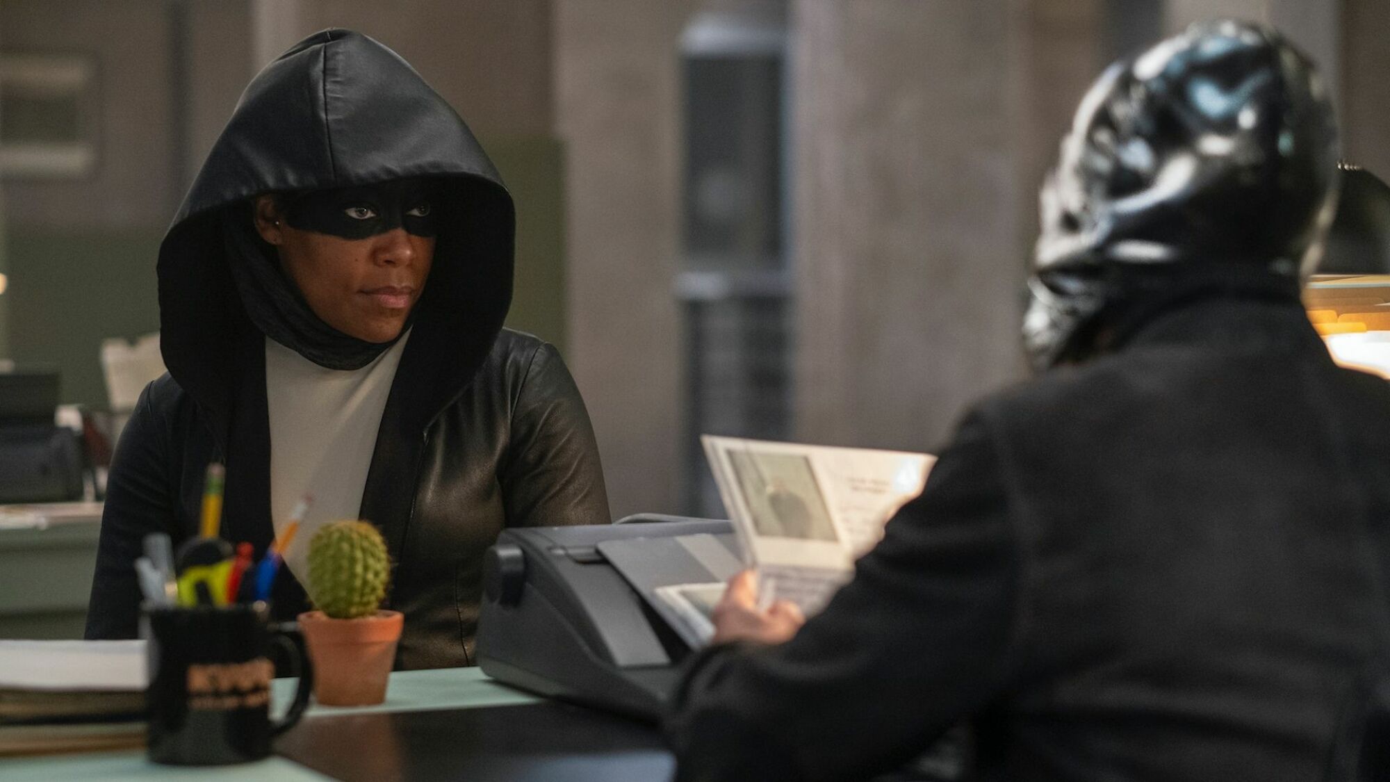 A woman in a mask and hood sits at a desk, staring at a person in a mask.