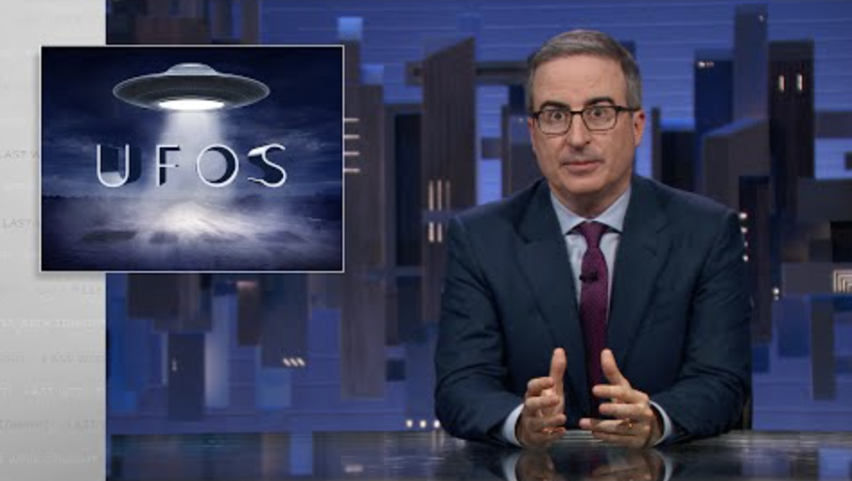 A man in a suit sits behind a talk show desk. In the top-left is an image of a UFO.