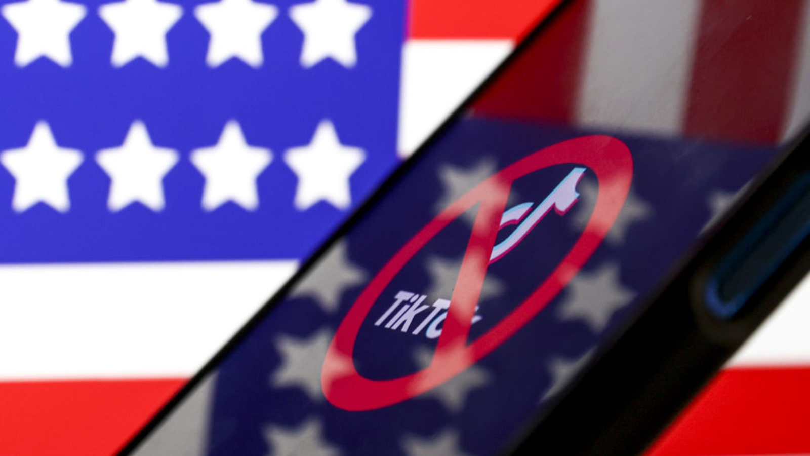 The TikTok logo with a cancellation symbol on top of it, displayed on a smartphone. It is in front of an American flag, which is also reflected on the smartphone screen.