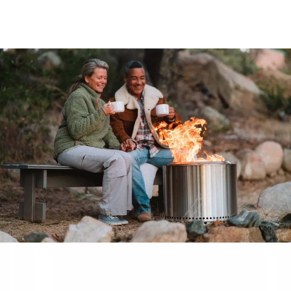 two people sit side by side on a bench behind a solo stove bonfire 2.0. They're both looking at the fire while smiling and both hold white mugs