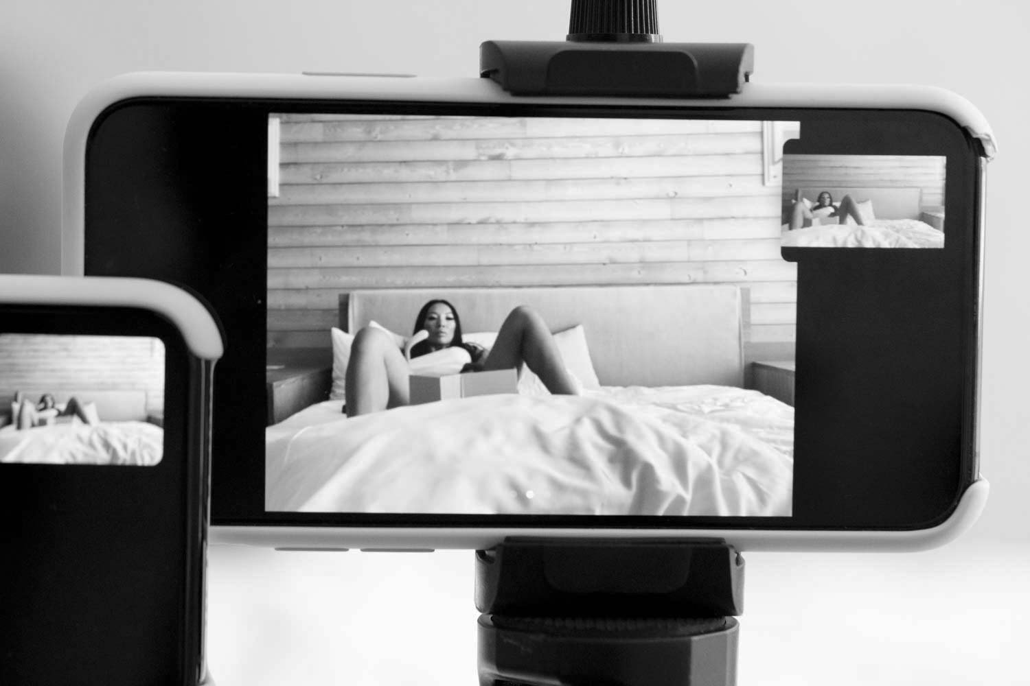 An iPhone sits on a tripod, filming a woman who lays with her legs open on a bed.