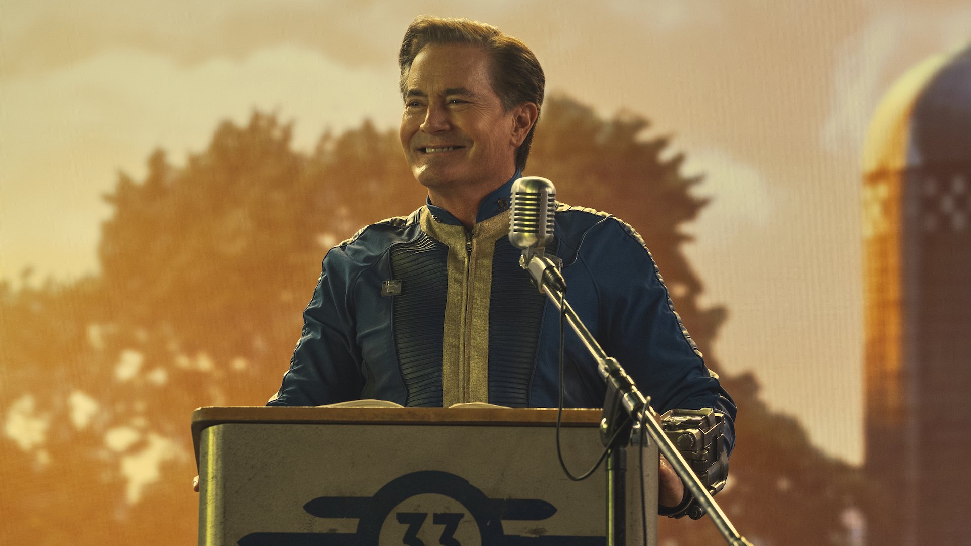 A smiling man in a blue and yellow jumpsuit smiles at a lectern giving a speech in 