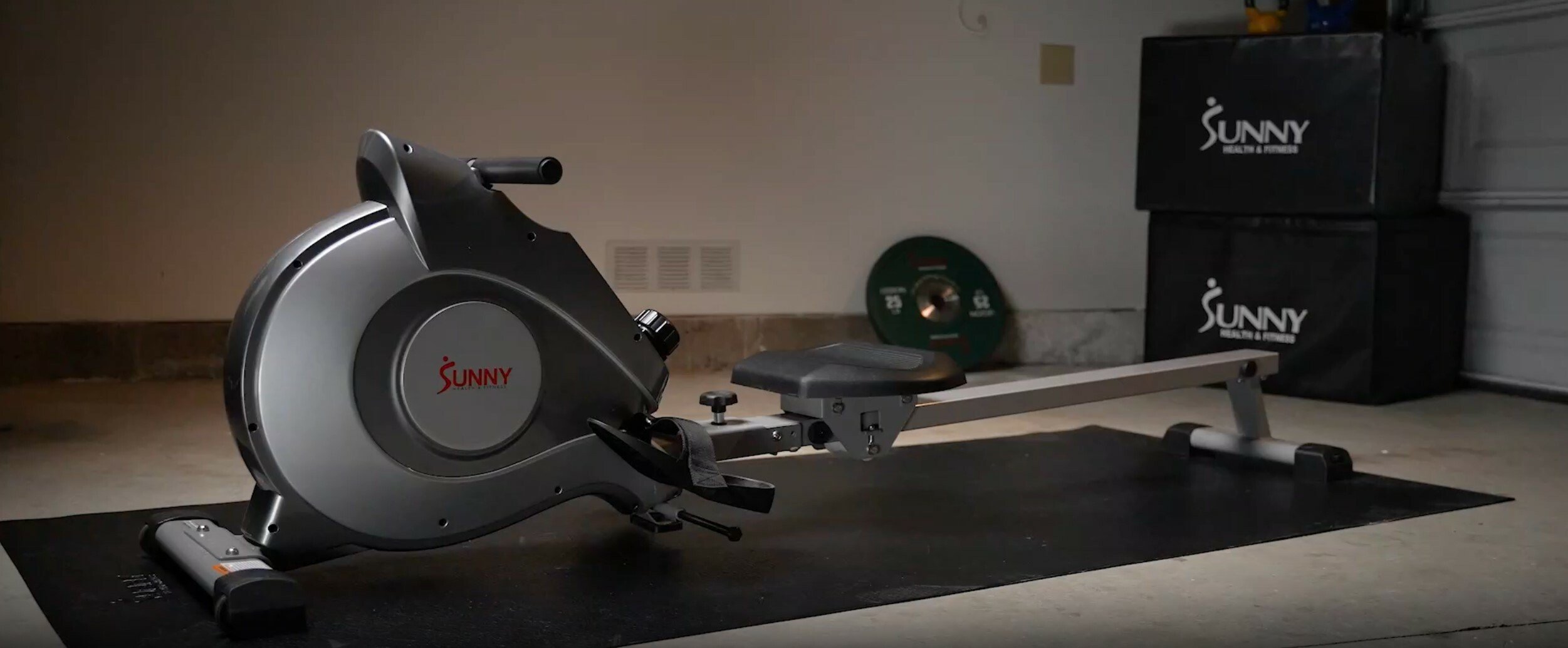 a sunny health rowing machine sits on a black mat on the floor with two large black boxes in the background that say sunny health
