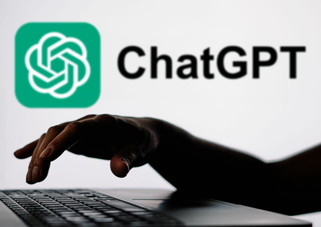 hand typing on a laptop in front of the ChatGPT logo on a wall
