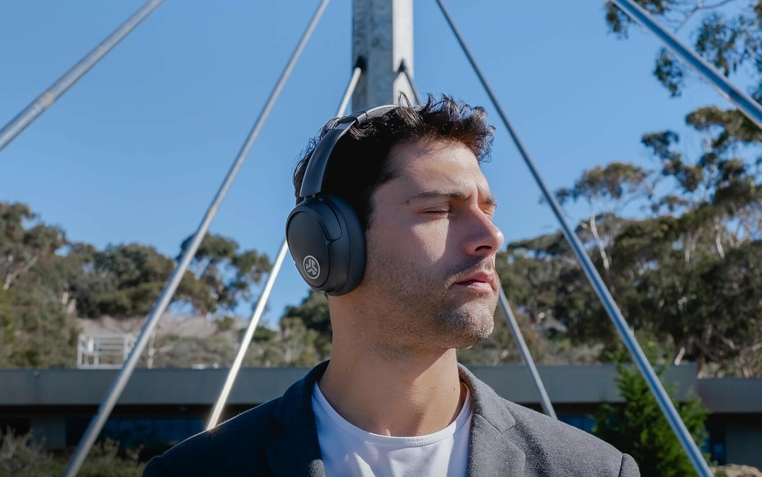 a male wears the jlab jbuds lux anc over-ear headphones while standing outside, facing the sun with his eyes closed 