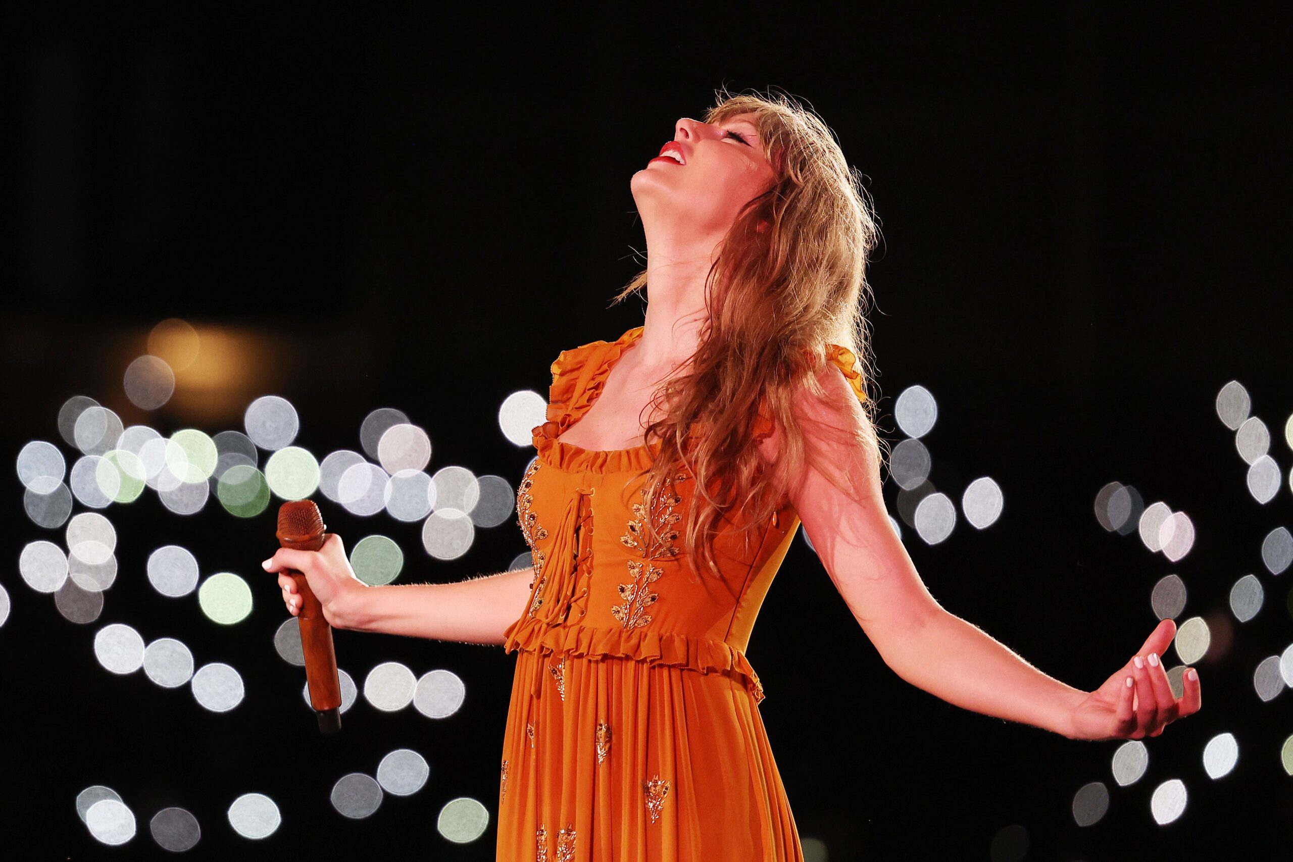 Taylor Swift in an orange dress looking up on stage. 