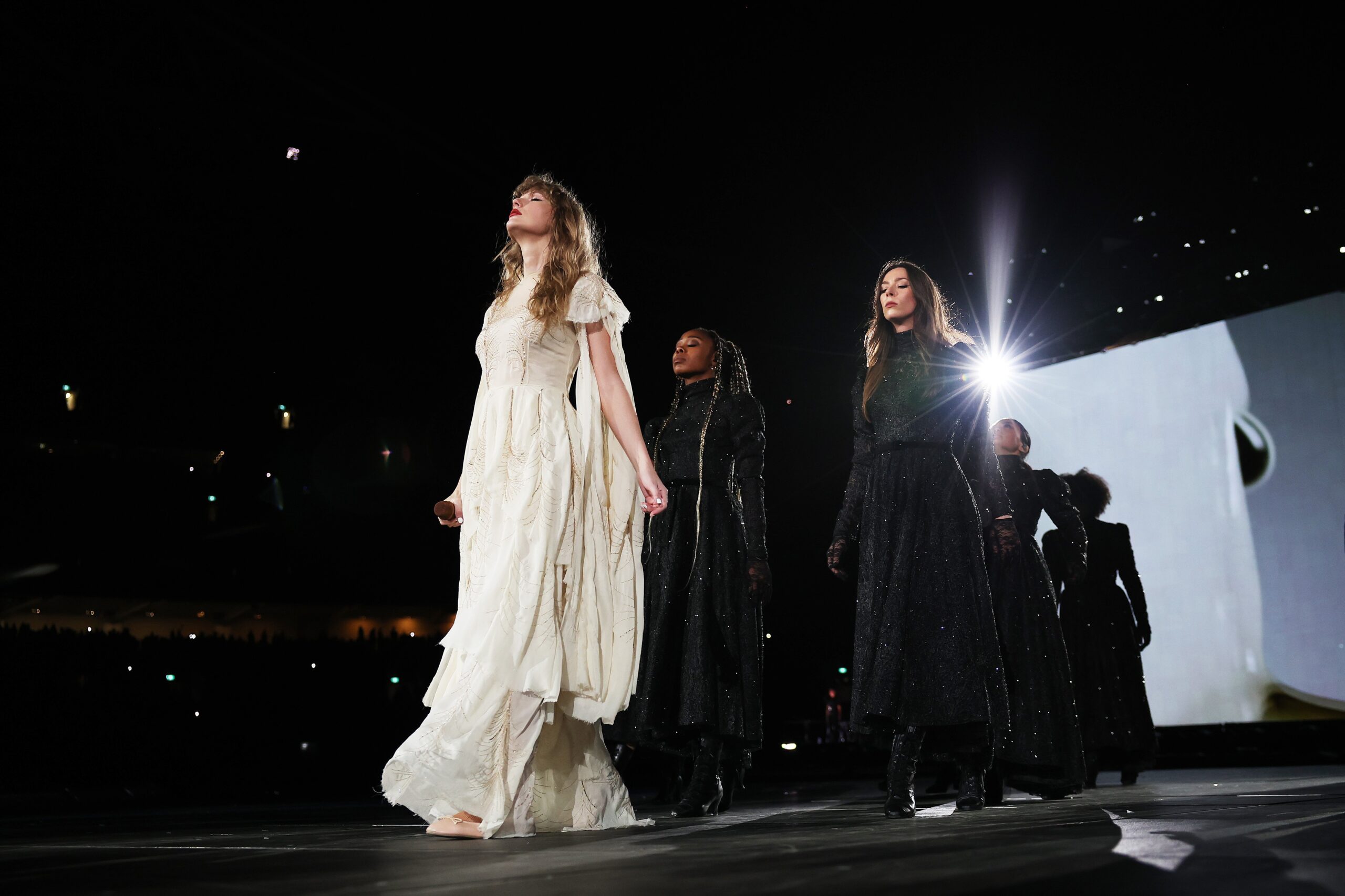 Taylor Swift performing in a white dress surrounded by back up dancers in black outfits creating a haunting image. 
