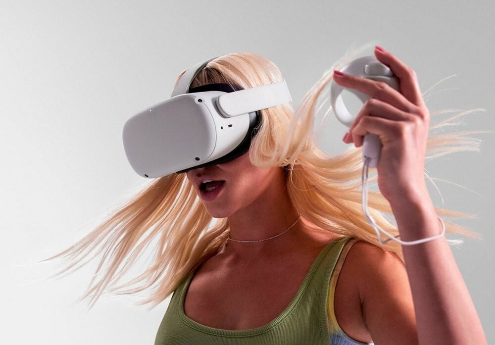 a person with long, blonde hair wears the meta quest 2 headset while using one of the hand tracking devices in her left hand