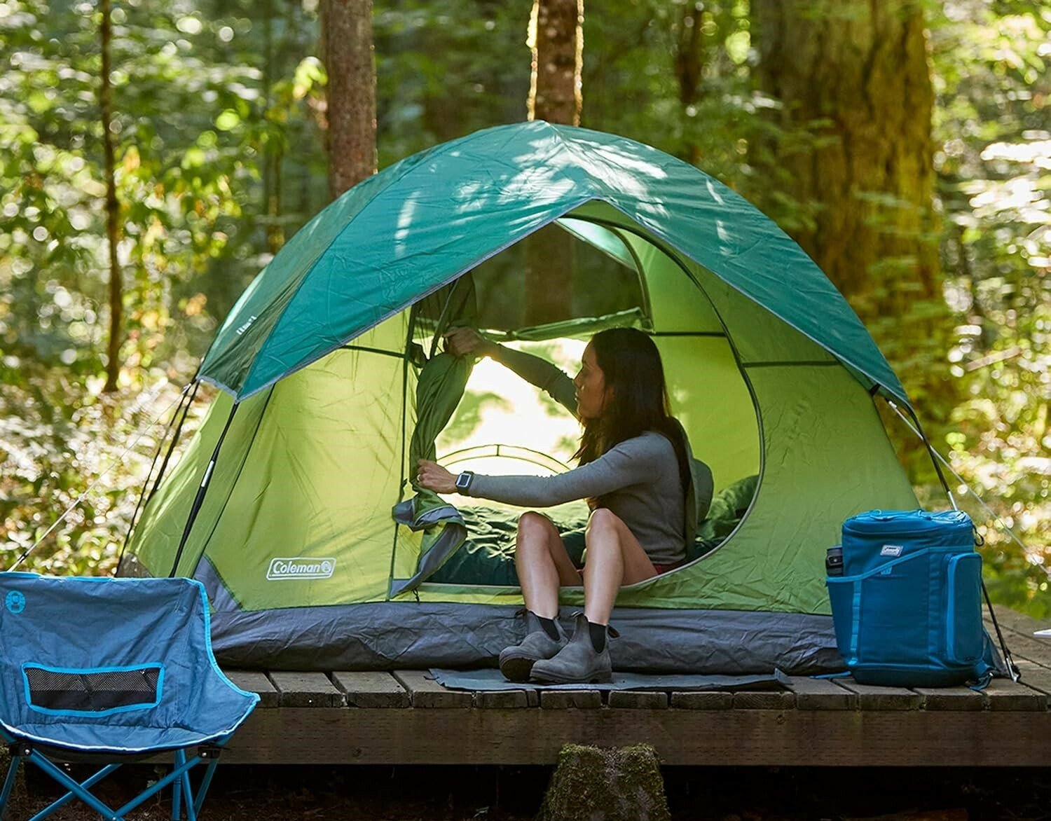 a person sits in a coleman tent that sits on a wooden platform in the woods