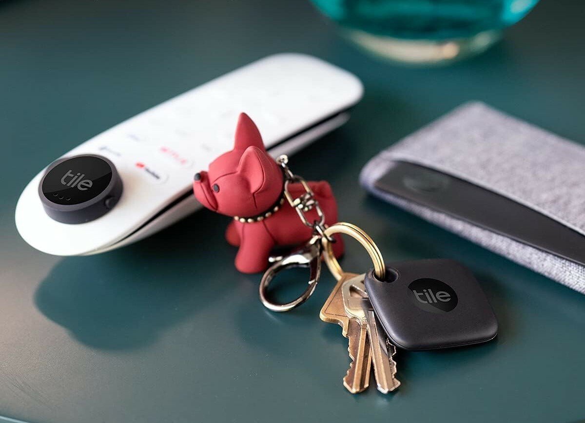 a side table includes a remote control, keys with a red dog keychain, and a wallet. All items have a tile bluetooth tracker attached.