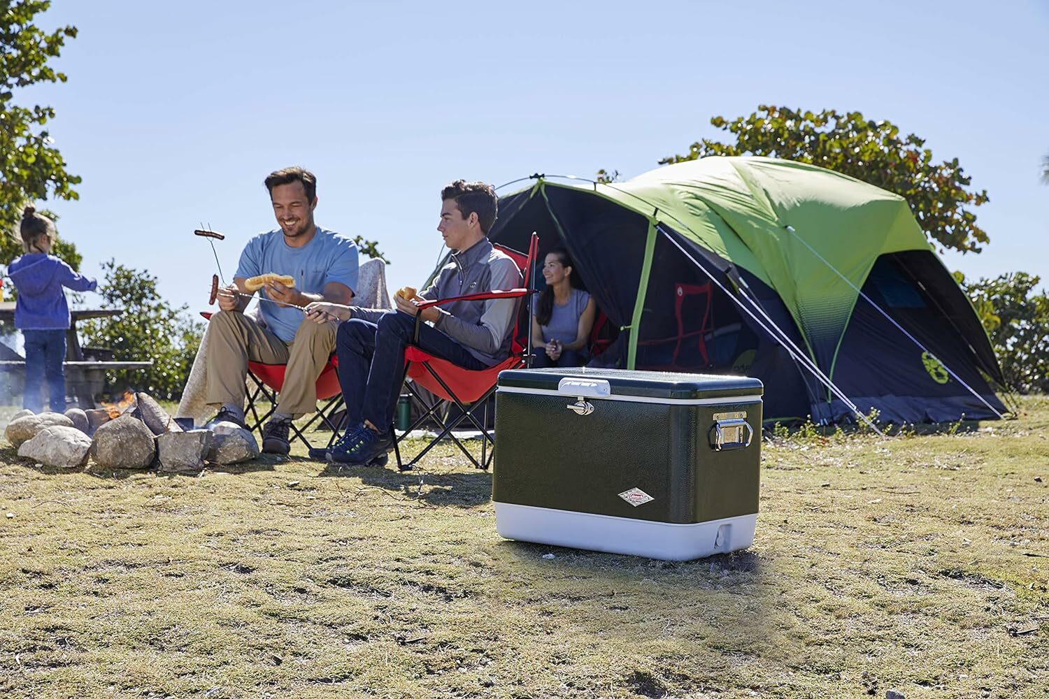 two people sit on red camping chair with skewers for hot dogs while a tent sits behind them and a cooler is in the foreground