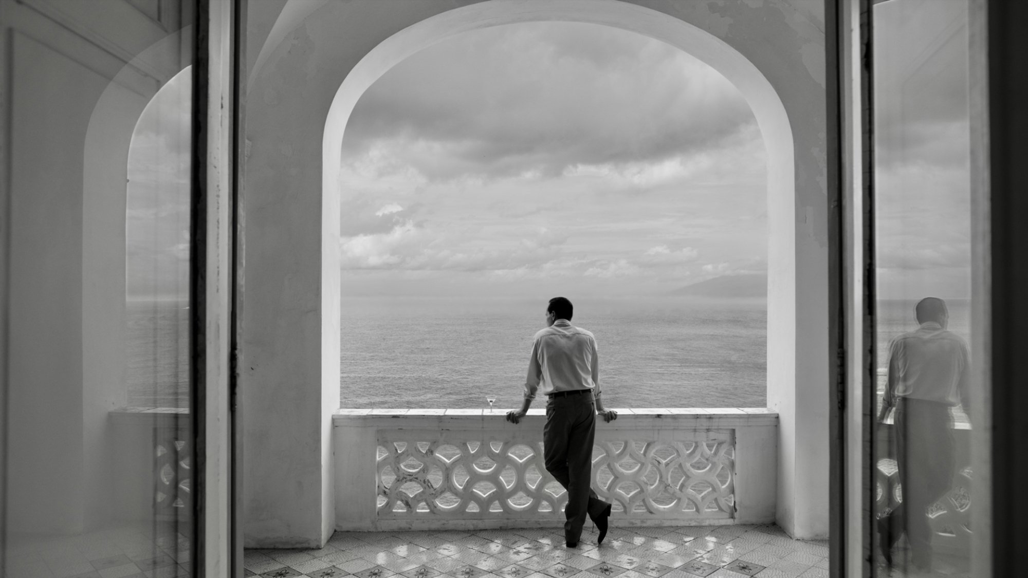 A man stands on a balcony overlooking the sea.