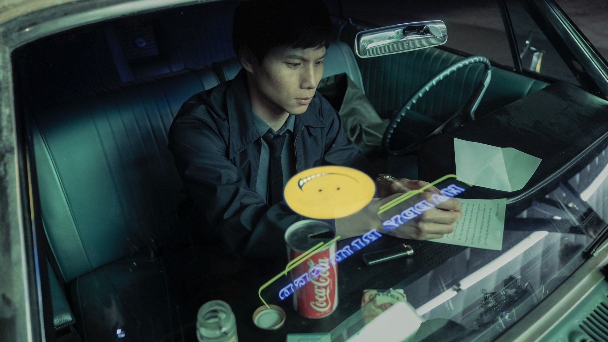 A man writing a letter in the front seat of a car; light from a yellow smiley face sign is reflected in the window. 