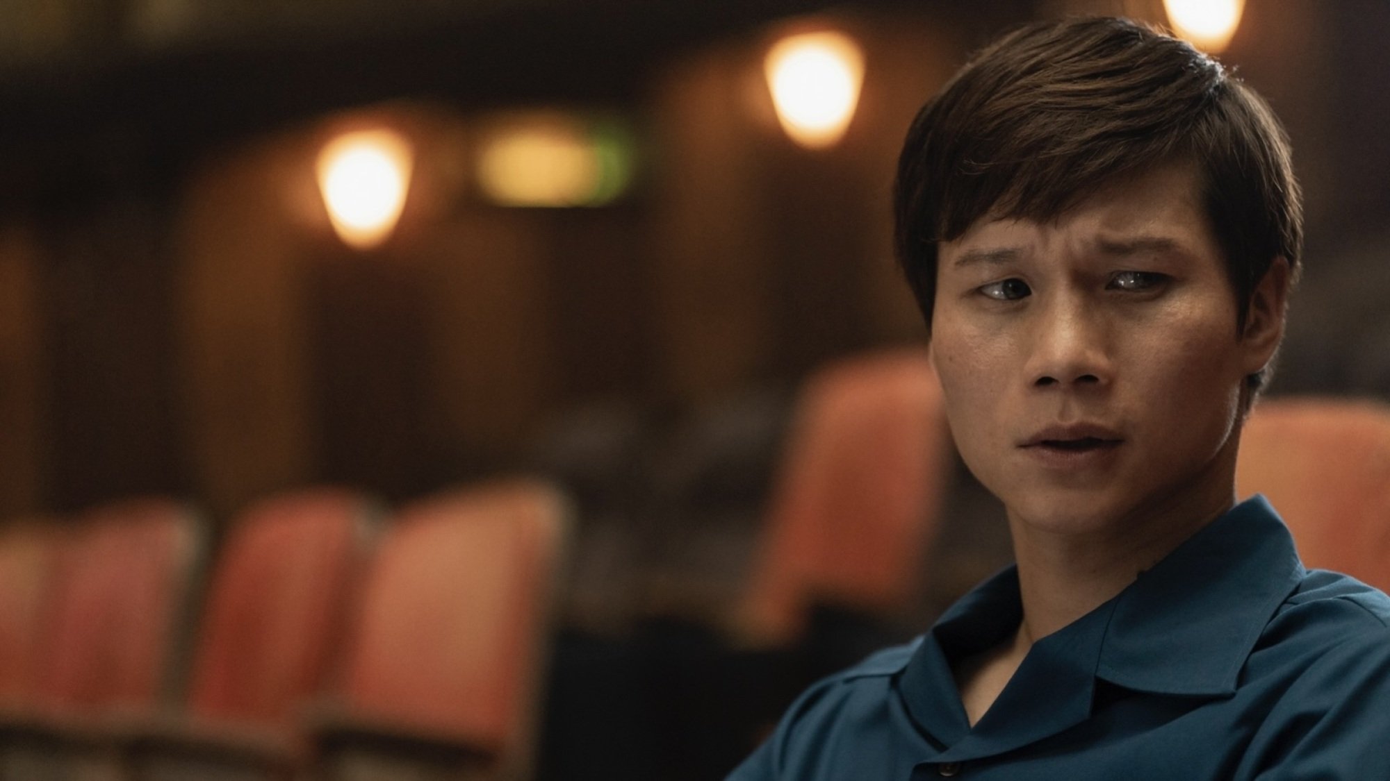 A man in a blue collared shirt sitting in a movie theater, looking anxious.