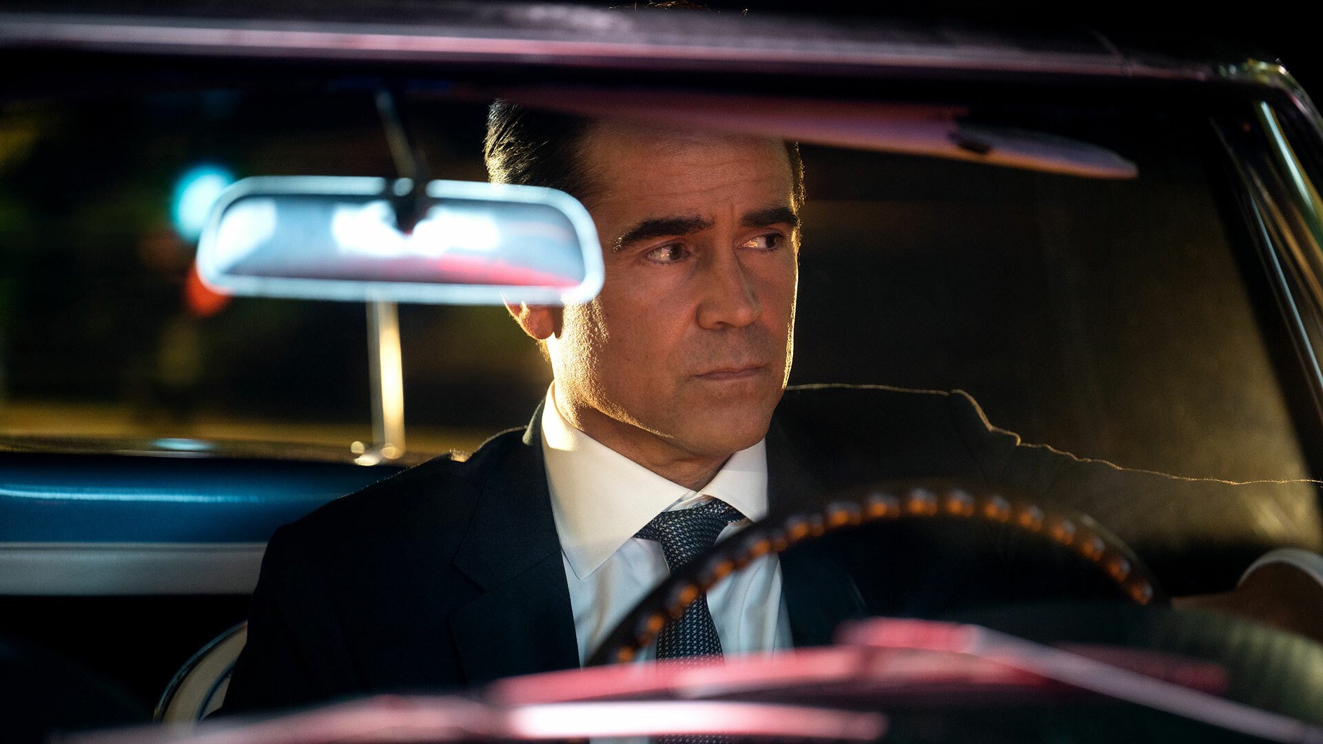 A man in a suit drives in an open top car at night.