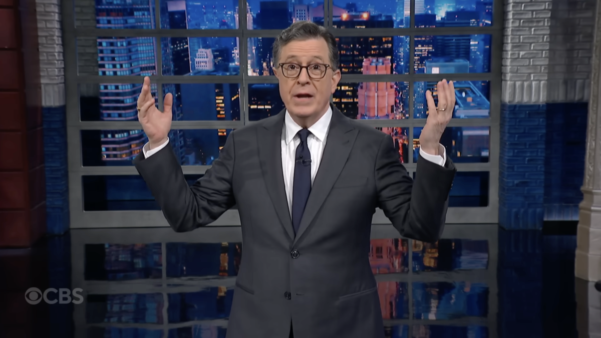 A man in a suit on a talk show stage gestures with his arms wide.