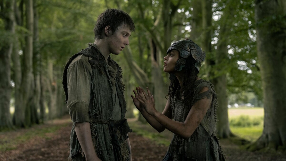 A boy and a girl stand on a tree-lined path. The girl wears a helmet of armour.