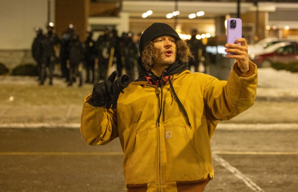 A man in winter gear with a purple iPhone in front of a line of masked officers.
