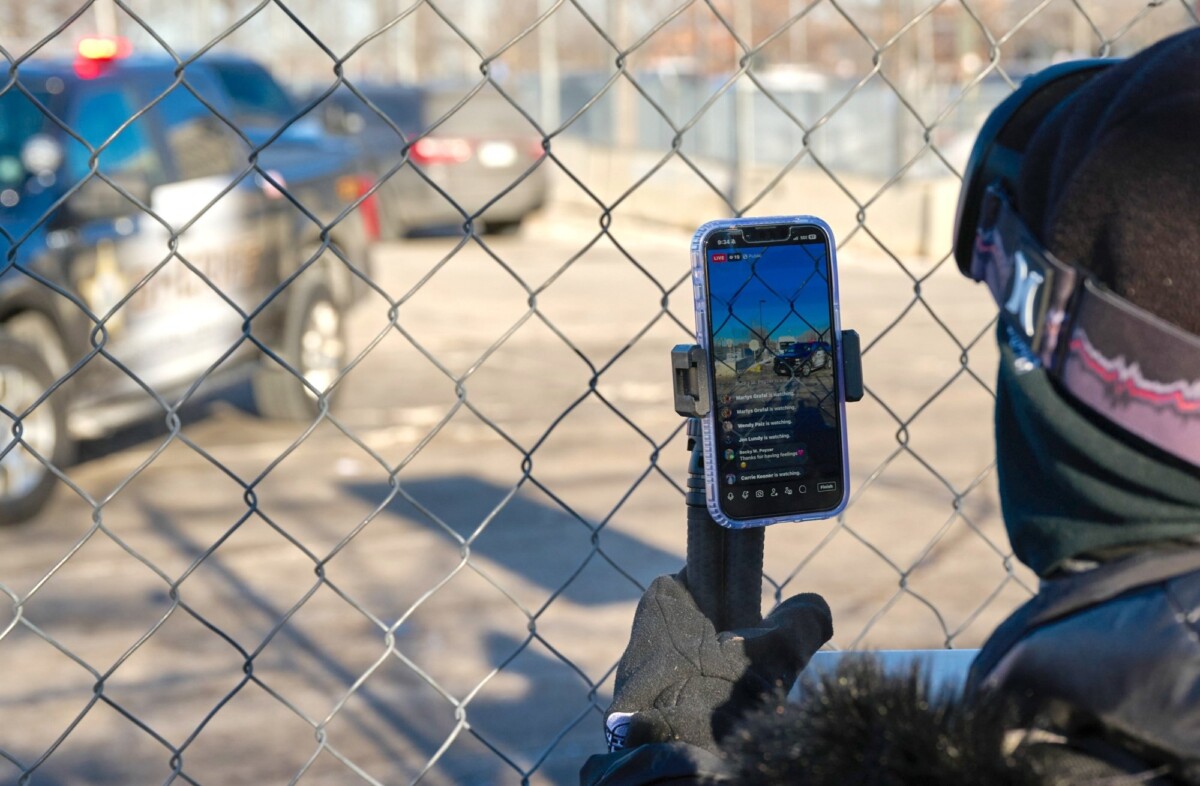 A masked observer with a phone showing video and comments behind a fence with official cars in front 