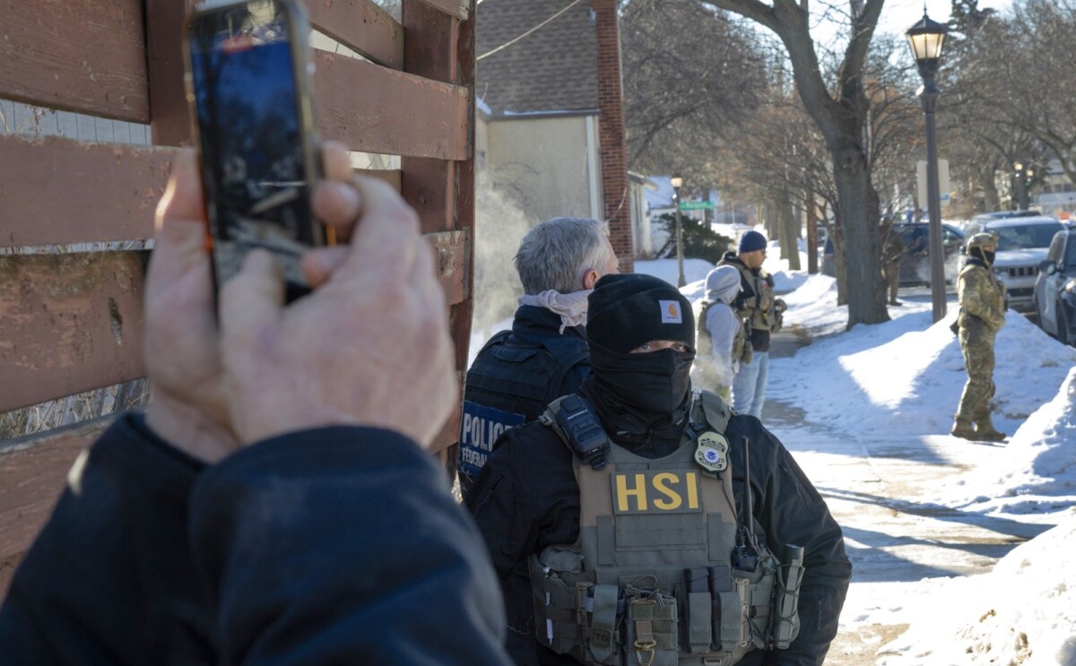 A man's hands holding a smartphone taking video of federal officers in masks in the snow. 