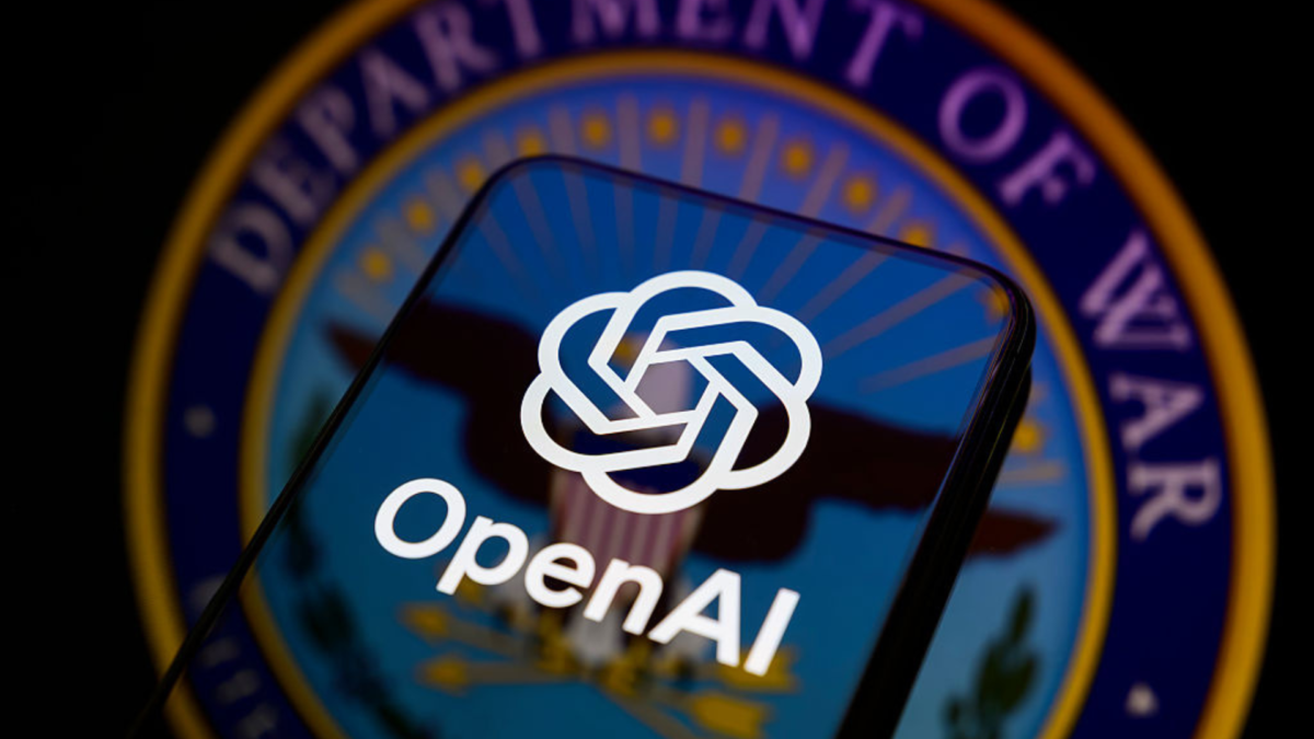 The OpenAI logo appears on the screen of a smartphone placed on a reflective surface where the seal of the United States Department of War (Department of Defense) is projected.